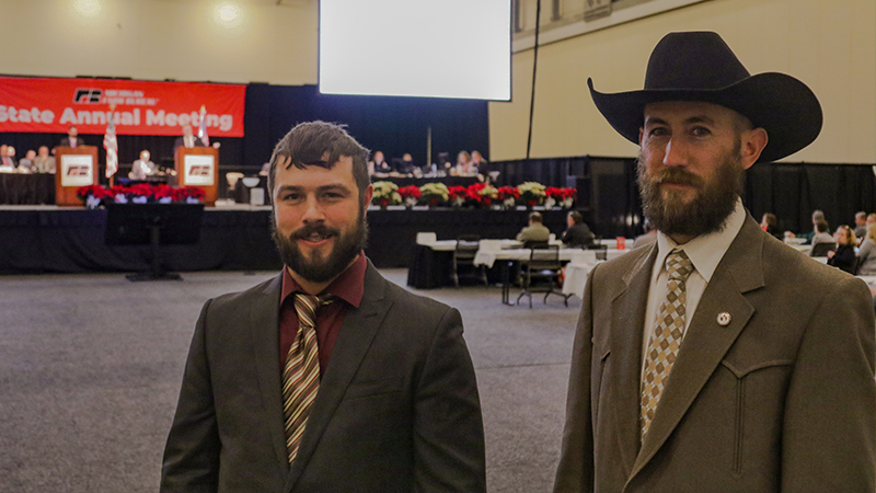 Two men in suits, one wearing a cowboy hat, standing on the floor of an exhibit hall while looking at the camera.