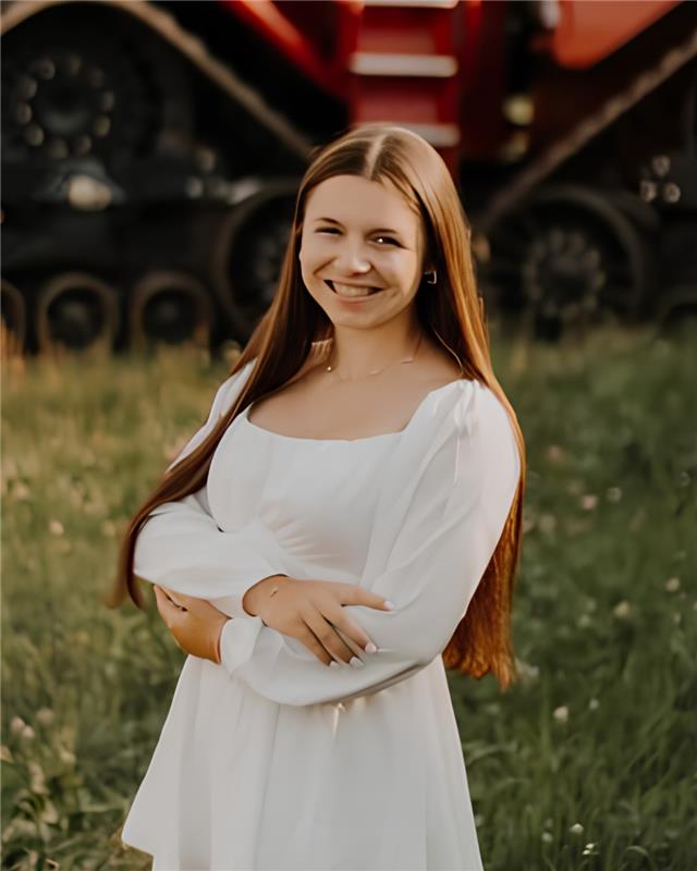 Nicole Neumann, scholarship recipient, standing in front of a tractor with arms crossed wearing white dress.