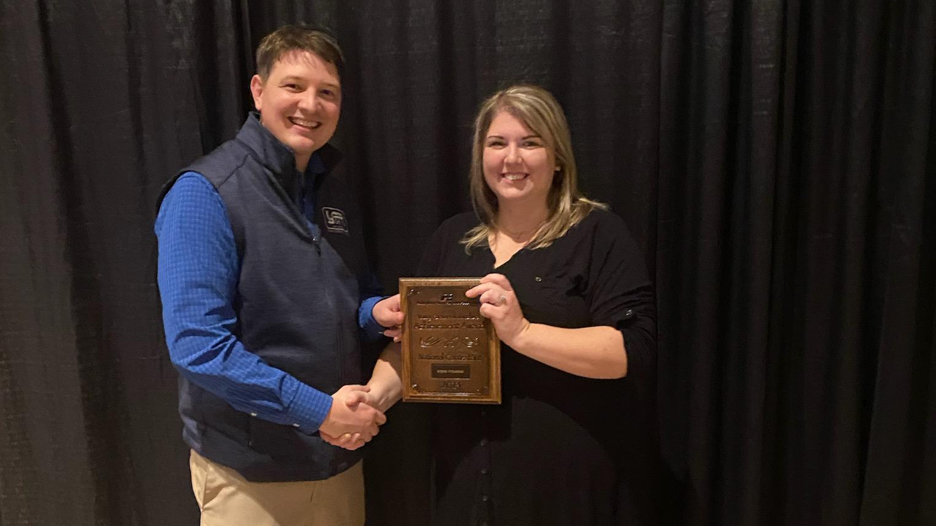 A man and a woman smiling at the camera and shaking hands as the man hands a plaque to the woman.