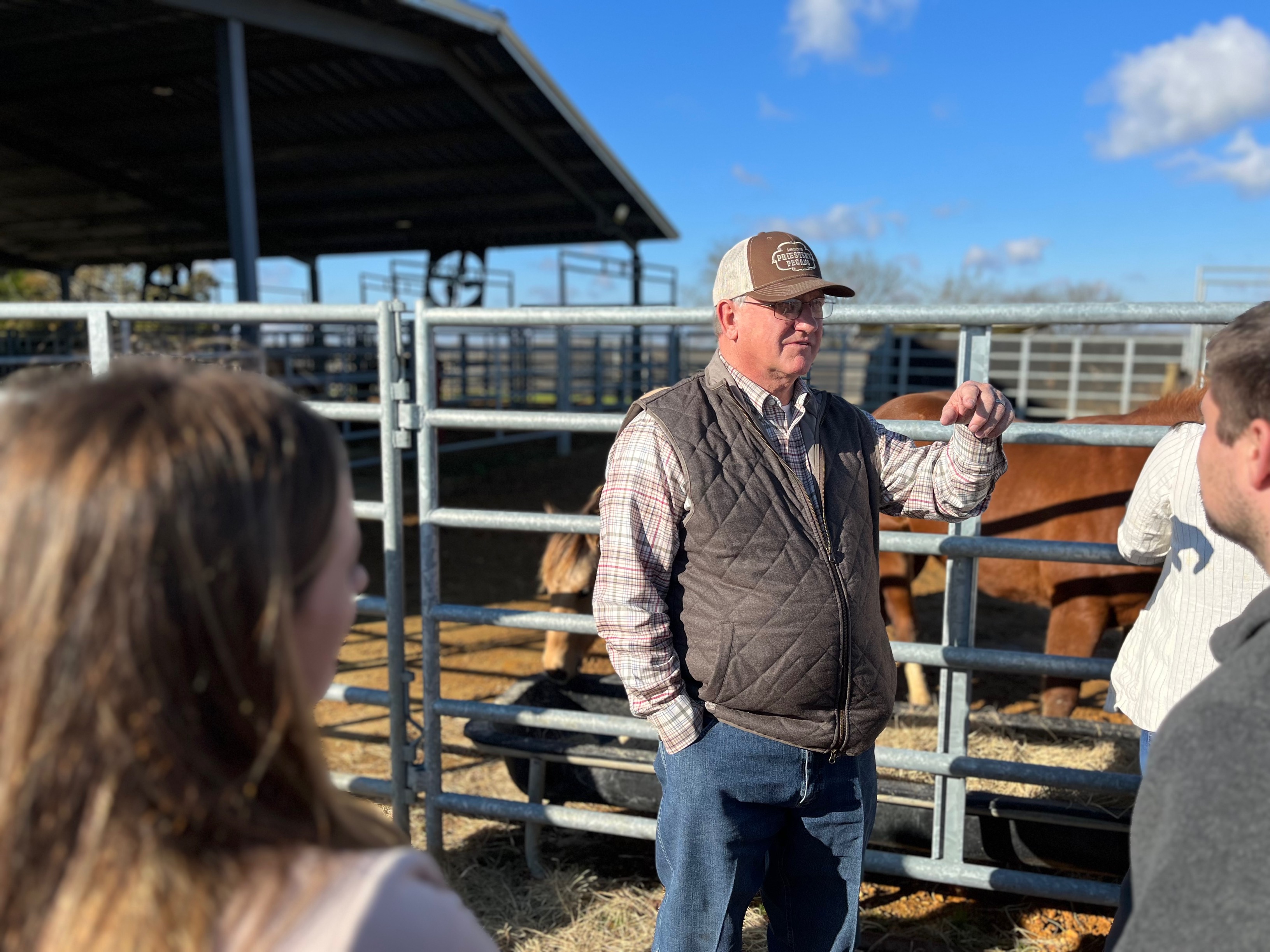 An older man in a hat and fest speaking to a group of young farmers in front of a cattle enclosure.