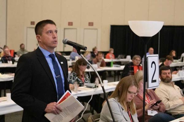 A man in a suit speaking into the microphone on the delegate floor of the 2022 MFB State Annual Meeting.
