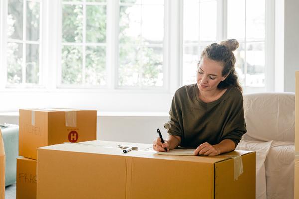 A woman sitting in a living room amongst a set of cardboard moving boxes while writing on a document on one of the boxes.