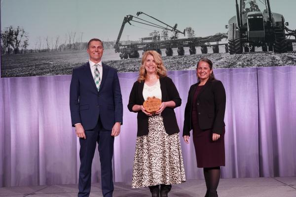 Portrait of Educator of the Year Award winner Katy Courtade smiling while holding her award next to MFB President Ben LaCross and MFB Board Member Alyssa Sanford on stage at the 2025 State Annual Meeting.