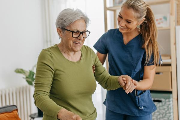 An in-home care assistant helping an elderly woman with a cane walk through her house.