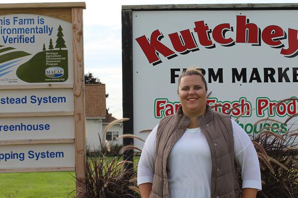 Portrait of Macomb County Farm Bureau president Amanda Kutchey smiling in front of a sign reading &quot;KUTCHEY'S FARM MARKET.&quot;