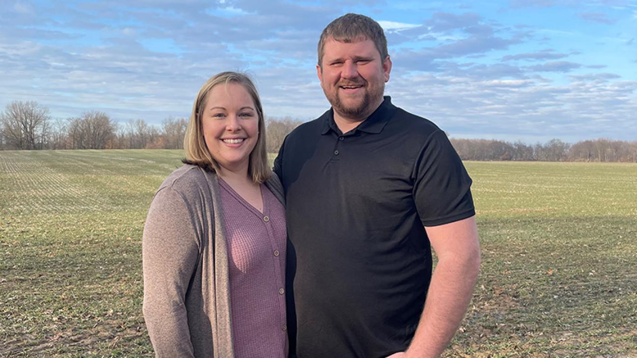 A male and female young farmer smiling at the camera while standing in a field.