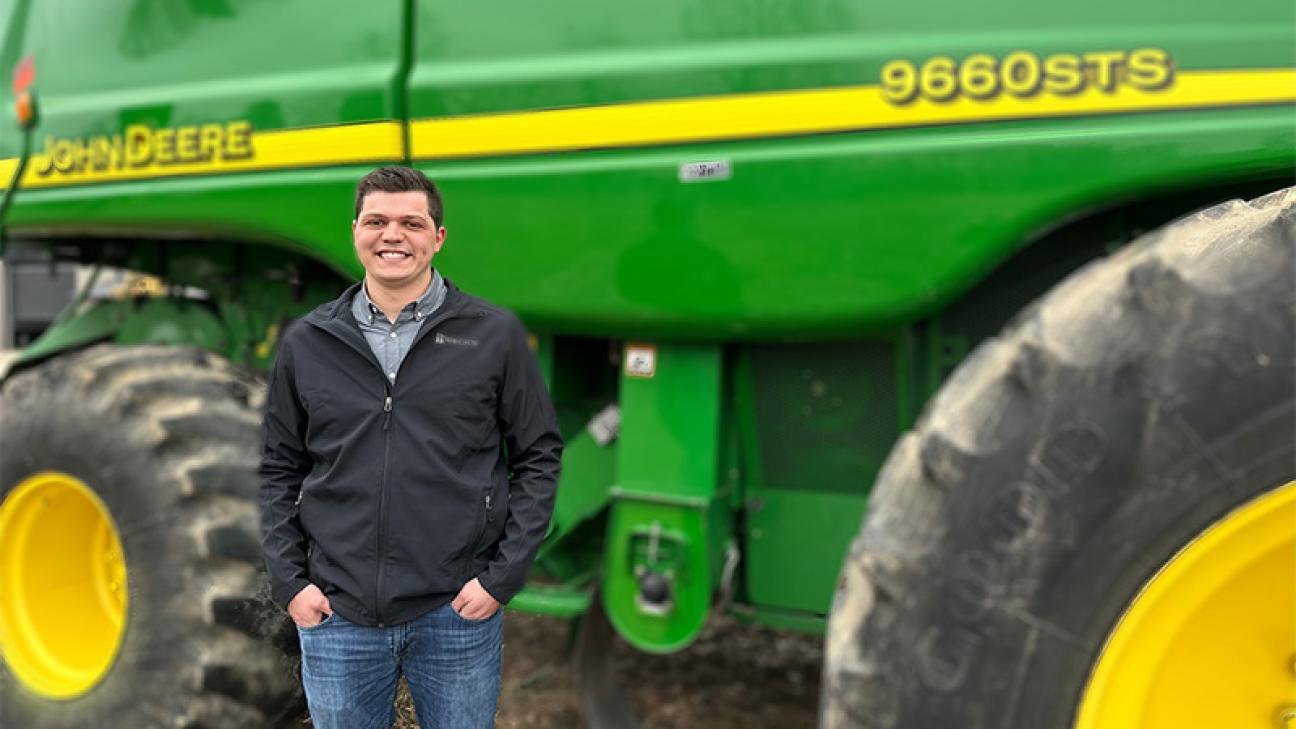 A male young farmer smiling at the camera while standing in front of a large green John Deere tractor.