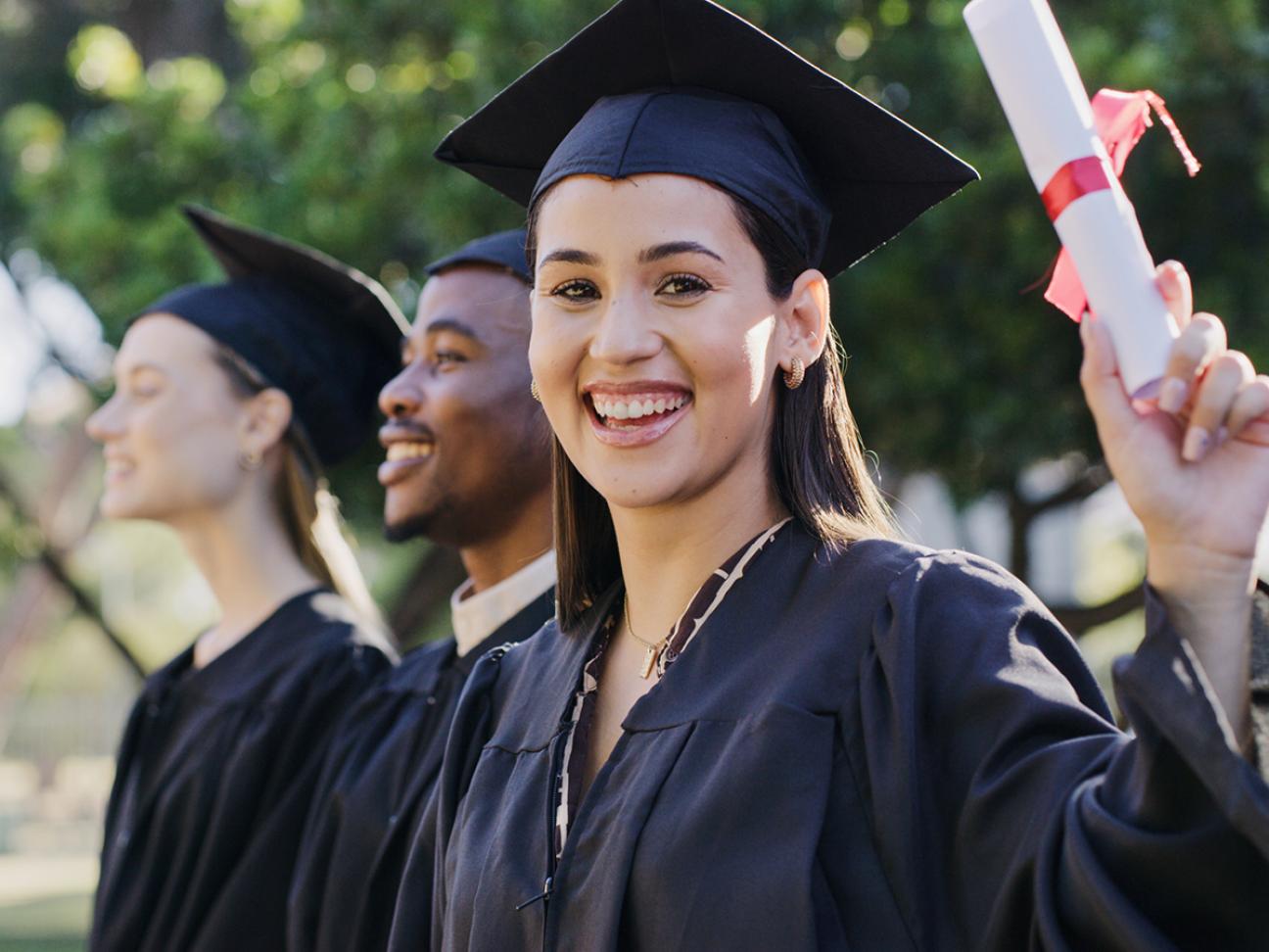 A student in a gown and graduation cap smiling at the camera while holding a diploma.