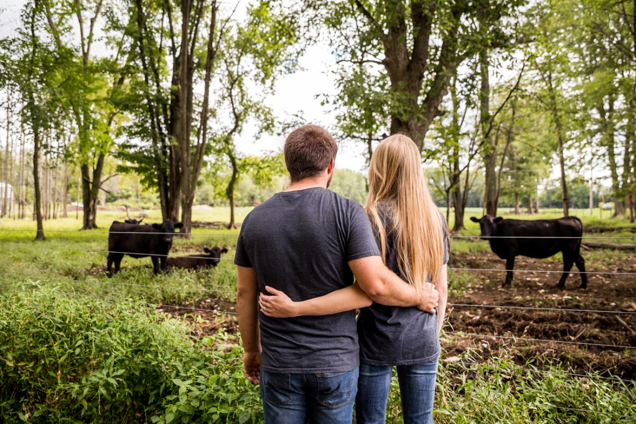 Rear view of two farmers with their arms wrapped around each other while looking upon a cattle herd in the woods.
