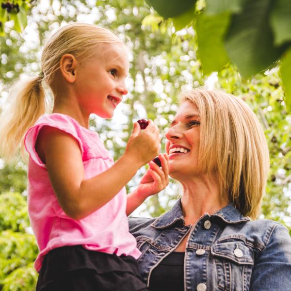 mother holds daughter outdoors while she eats a cherry