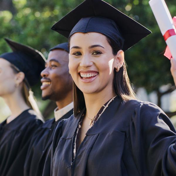 A student in a gown and graduation cap smiling at the camera while holding a diploma.