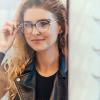 Woman in a leather jacket smiling as she tries on glasses and looks in the mirror