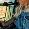 View from the cabin of a large tractor operating in a field of an older farmer in bib overalls operating the machine.